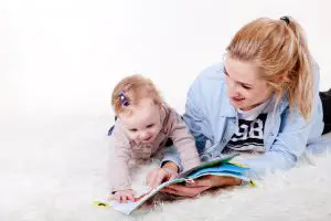 Toddler avoiding sleep time, with mom reading a book in How many hours should a toddler sleep